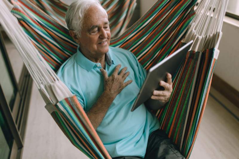 A senior man relaxing on a hammock and smiling at his tablet, representing the financial benefits of tax credits for seniors and how they can improve financial stability.