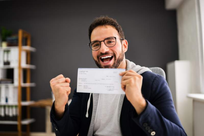 A man excitedly holds up his paycheck with one hand while pumping his fist, celebrating his bonus after taxes are withheld.