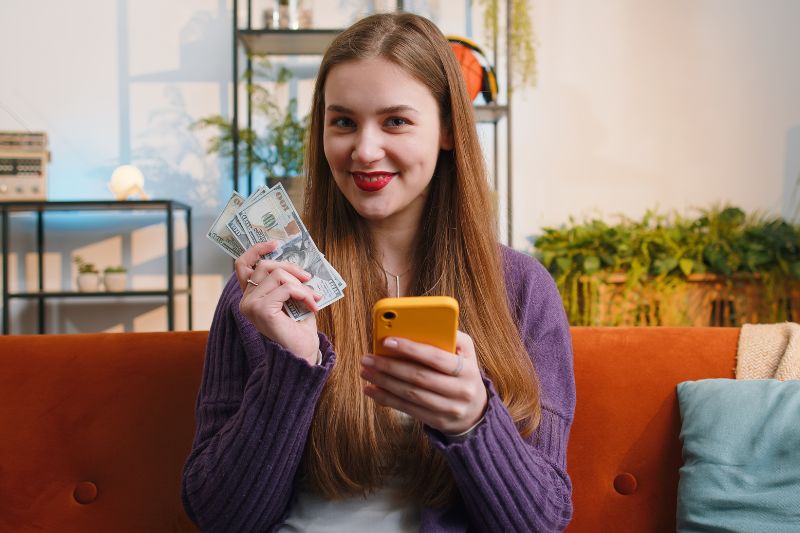 A smiling woman holding money in one hand and a phone in the other, sitting in her living room, symbolizing tax filing without a W-2.