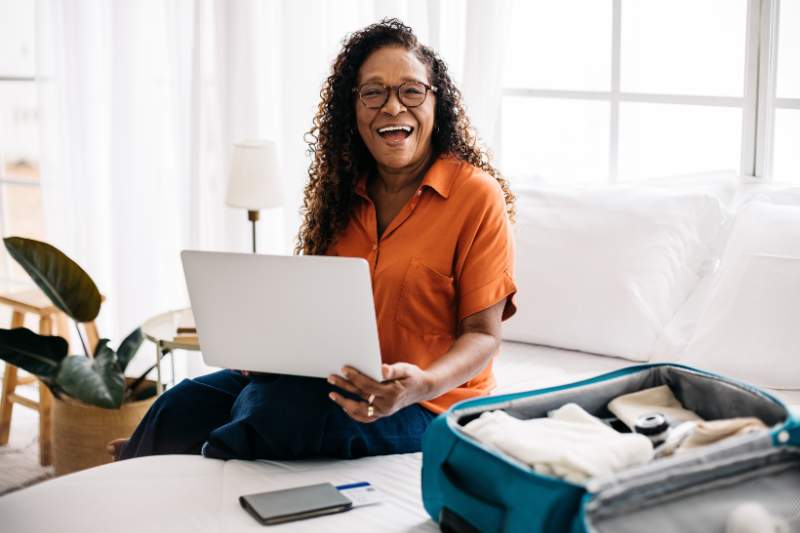 Happy woman sitting on her bed with a suitcase and laptop, ready to book her next flight with tips for saving money on airfare.