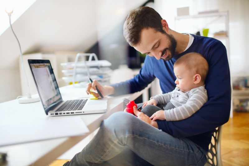 A dad sitting at his home office desk with his baby on his lap, smiling, symbolizing the Head of Household filing status for tax purposes.