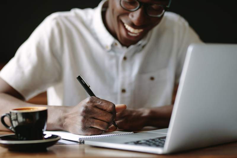 Man smiling at his laptop while taking notes on tax deductions he can claim for maximum savings.