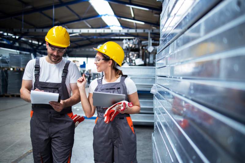 Two factory workers discussing the difference between 1099 and W-2 forms, with one holding a notepad and the other a tablet.