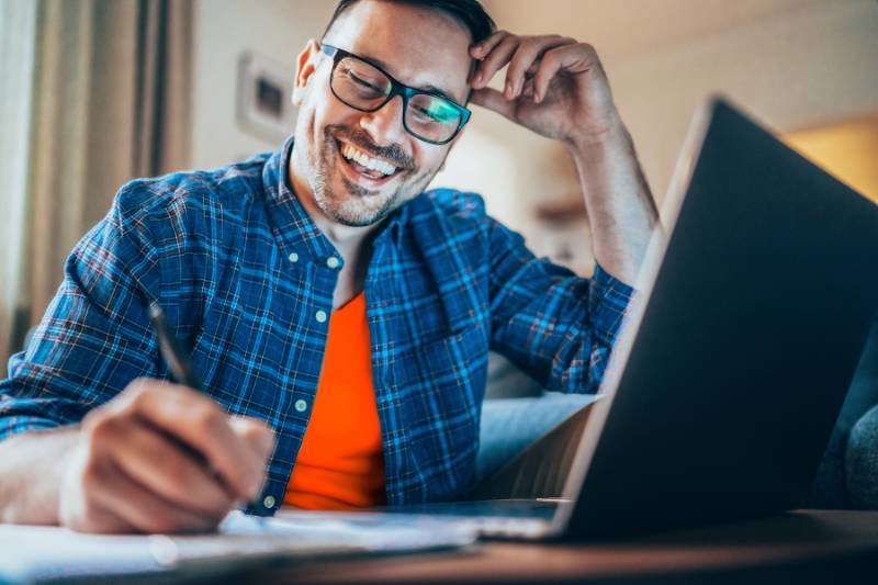 Smiling man taking notes on a notepad while using his laptop, representing the concept of tax write-offs and financial planning.