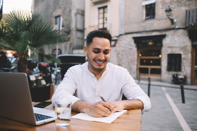 A cheerful man working on his laptop at an outdoor cafe, taking notes on above-the-line deductions to maximize tax savings.