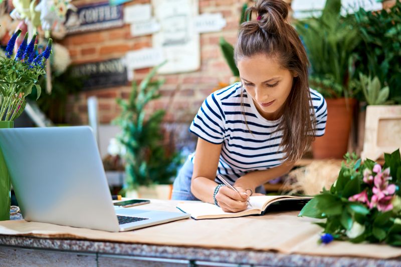 A florist working as a 1099 independent contractor, writing in her notebook while managing her business finances on a laptop, preparing for tax filing.